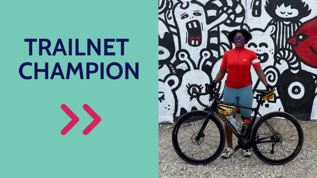 On the left side in blue against a teal background it says Trailnet Champion. To the right is a picture of Dorris Scott, the October Trailnet Champion; she is a Black woman wearing a red bike jersey, standing behind her black bike.